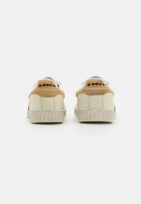 White leather sneakers with beige suede accents, a gray rubber sole, and a logo on the heel. Features perforations for breathability.