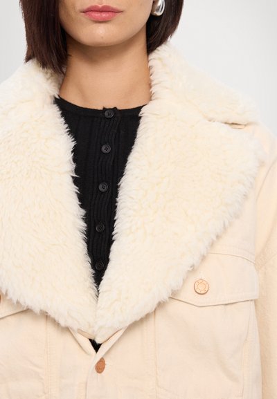 Chaqueta crema con un cuello blanco esponjoso, cierre de botones y una blusa negra de canalé debajo. Presenta una textura de tela suave.