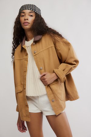 Frau mit langen lockigen Haaren trägt ein gemustertes Stirnband, eine übergroße braune Jacke, einen cremefarbenen Strickpullover und weiße Shorts vor einem schlichten Hintergrund.