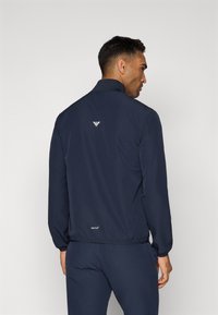 Ναυτικό μπλε αδιάβροχη ζακέτα με ψηλό γιακά, ελαστικές μανσέτες και λεπτή γραμμή. Περιλαμβάνει ένα μικρό λογότυπο στον λαιμό και το στρίφωμα. Λείο υλικό.