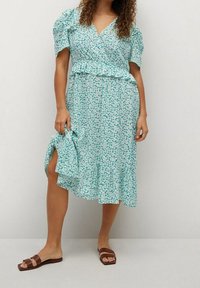 Vestido envolvente floral en tonos azul verdoso, con mangas cortas abullonadas, cintura fruncida y falda a la altura de la rodilla. Combinado con sandalias marrones de tipo slide.