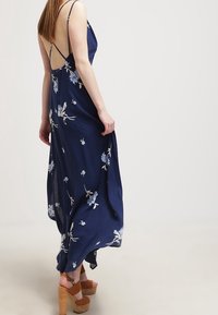Vrouw in een marineblauwe bloemenmaxi-jurk met dunne bandjes en een open rug, gecombineerd met tan hoge sandalen met enkelbandjes.