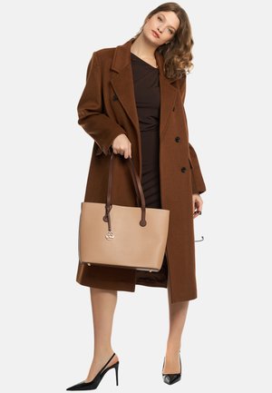 Brun uld dobbeltradet frakke, mørk kjole, sorte lakhæle, holder en beige tote med brune stropper og et logoaccent.