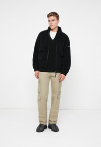 Ung man står klädd i en svart zip-up fleecejacka, svart V-ringad tröja, beiga cargobyxor och svarta sneakers mot en vit bakgrund.