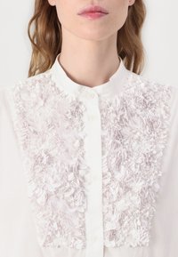 Blouse blanche avec un design texturé en applique florale de couleur rose doux sur le devant, dotée d'un col et d'une fermeture à boutons.