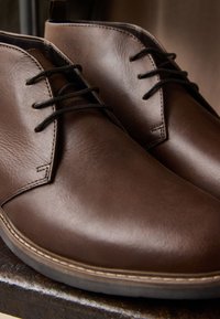 Hnedé kožené topánky s hladkou textúrou a zaoblenou špičkou, vybavené čiernymi šnúrkami a jemnými švami pozdĺž podrážky.