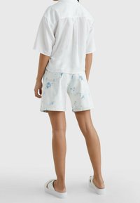 Maglietta bianca a maniche corte, pantaloni corti blu chiaro con tie-dye, sandali bianchi senza chiusura. La vista posteriore mostra la piega della maglietta e le tasche dei pantaloni.