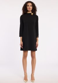 Robe noire en ligne A avec un col montant, des manches trois-quarts et un accent en forme de fleur dorée sur le col. Texture de tissu lisse.