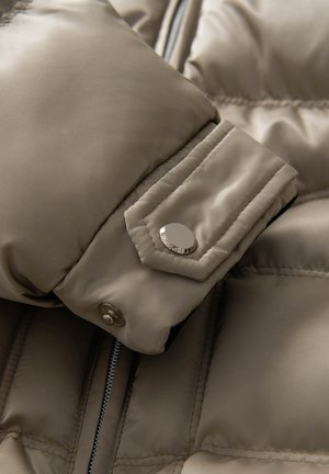 Beige puffjacka i detalj med satinfinish, framträdande sömmar och en silverspänneknapp på ärmen. Synlig dragkedja på sidan.