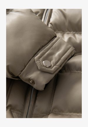 Beige Puffermantel-Detail mit satinierter Oberfläche, auffälliger Naht und silbernem Druckknopf am Bündchen. Sichtbarer Reißverschluss an der Seite.