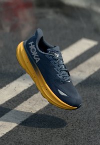 Hoka løpesko med et navy mesh-overdel og en tykk gul såle, designet for demping, med fremtredende logo og teksturert overflate.