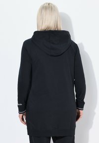 Henkilö, jolla on suorat vaaleat hiukset, on pukeutunut pitkähihaiseen mustaan huppariin ja seisoo kasvot kohti yksinkertaista valkoista seinää.