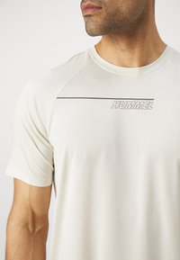 Light beige athletic shirt with a ribbed texture. Features a round neckline and short sleeves. "HUMMEL" logo in dark gray across the chest.