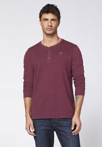 Bordeauxrotes Langarm-Henley-Shirt aus Baumwolle, mit einem Drei-Knopf-Verschluss und dezentem Logo-Detail auf der Brust. Getragen mit dunklen Jeans.