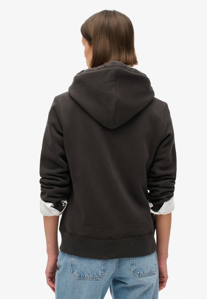 Dunkelgrauer Hoodie mit einer großen Kapuze, gerippten Bündchen und Saum. Getragen über einem weißen Shirt mit hochgerollten Ärmeln, kombiniert mit blauen Jeans.