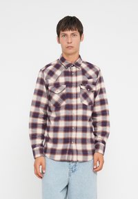 Camisa de franela a cuadros en crema, rojo y azul marino. Presenta un cuello puntiagudo, dos bolsillos en el pecho y cierre de botones. Textura suave con un corte relajado.