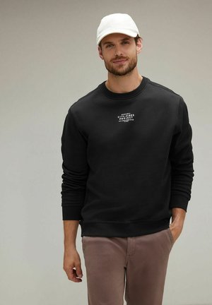 Schwarzer Sweatshirt mit Rundhalsausschnitt und Rippenbündchen, auf dessen Vorderseite weißer Grafiktext zu sehen ist. Getragen mit hellen Hosen.
