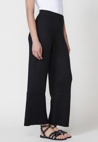 Pantalons larges noirs à côtes avec une taille élastique, présentant une surface texturée. Portés avec des sandales noires à lanières, vue de côté montrée.