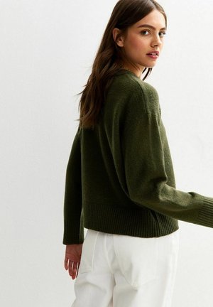 Suéter corto de un color verde oliva, con ribetes en el dobladillo y las mangas, que presenta una textura suave. Modelo visto desde atrás, que lleva pantalones blancos.