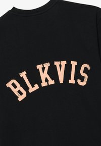 Camiseta negra con letras curvas y desvanecidas en melocotón que dicen "BLKVIS" en la parte de atrás. Elaborada con una tela suave y texturizada.