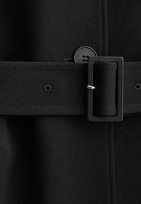 Cinturón de tela negra con hebilla rectangular abrochada sobre abrigo negro a juego con textura, con detalles visibles de botones y costuras.