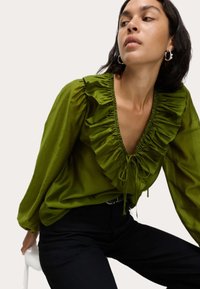 Groene blouse met gerimpelde halslijn, lange mouwen en strikdetail. Gladde stof, losse pasvorm, gecombineerd met donkere broeken.
