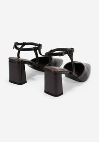 Zapatos de tacón de color marrón oscuro con puntera afilada y tacón grueso. Cuentan con una correa en el tobillo con hebilla y una textura de cuero suave.