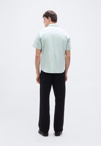 Άνδρας που στέκεται πλάτη, φοράει ανοιχτό μπλε πουκάμισο με κοντά μανίκια, μαύρο παντελόνι και μαύρα παπούτσια σε λευκό φόντο.