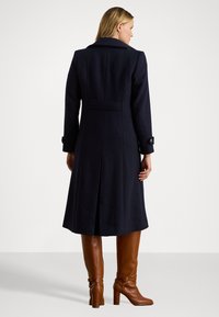 Abrigo de lana marino con corte entallado, que presenta un cuello muesca, bolsillos laterales y puños abotonados, combinado con botas de cuero marrón hasta la rodilla.