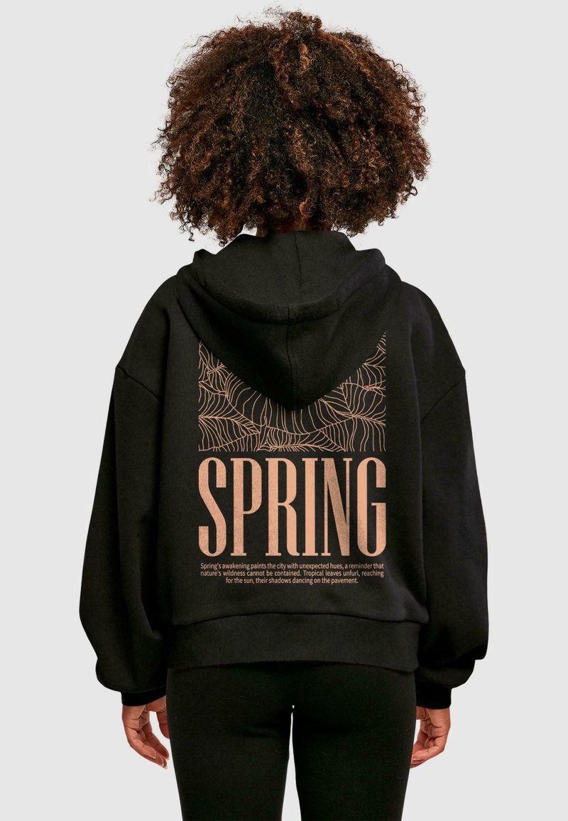 Sort hættetrøje med stort trykt tekst "SPRING" i fed skrift og et detaljeret bladmønster på ryggen. Blødt materiale, oversized pasform.