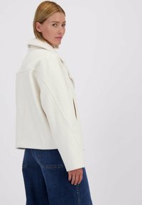 Vrouw met kort blond haar draagt een witte wollen jas en blauwe jeans, staat in profiel tegen een effen witte achtergrond.