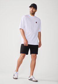 White t-shirt with black logo, paired with black shorts and white sneakers featuring black stripes. Short sleeves and relaxed fit.