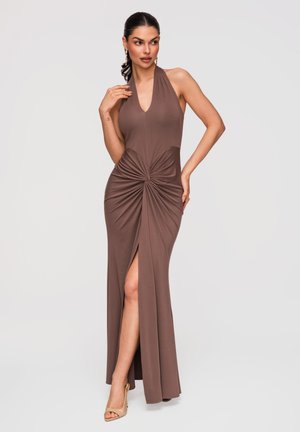 Vrouw draagt mouwloze bruine halterjurk met knoopdetail aan de voorkant en een hoge split, gecombineerd met beige open-toe hakken, poserend tegen een effen achtergrond.