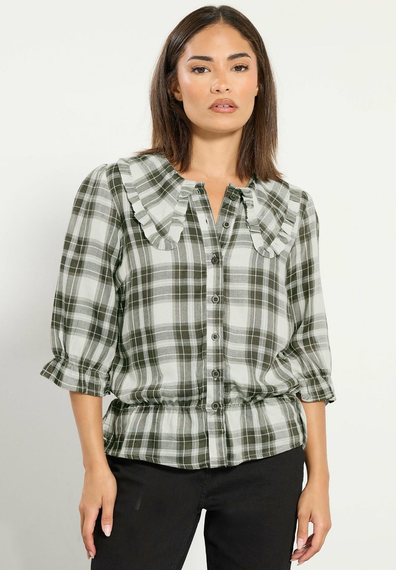 Mujer con blusa de cuadros verdes y blancos con cuello y mangas con volantes, de pie frente a un fondo claro y liso.
