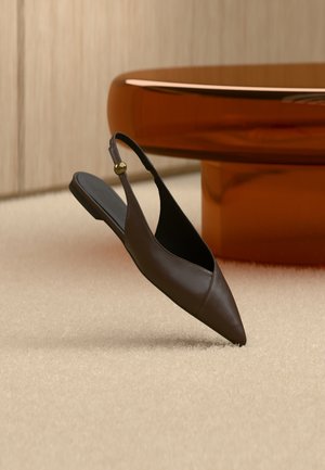 Chaussure plate en cuir marron foncé à bout pointu avec bride arrière et boucle dorée, posée sur un tapis beige près d'une table ronde en bois.