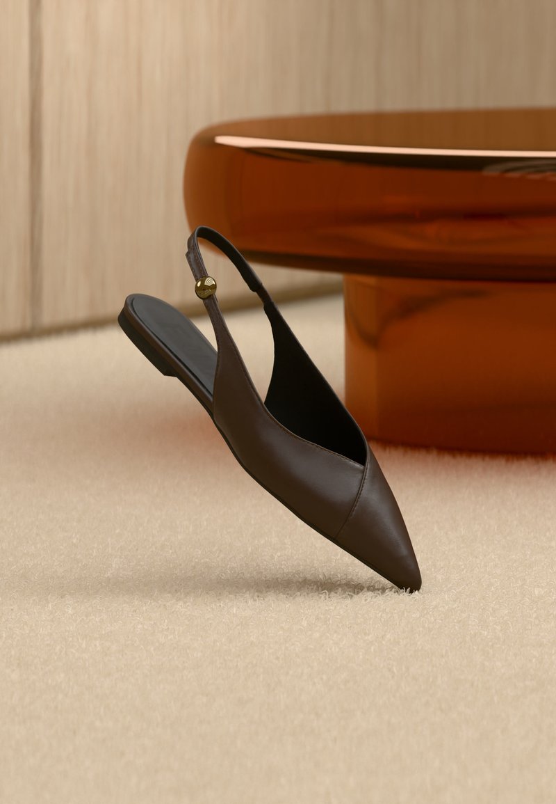 Σκούρο καφέ δερμάτινο γυναικείο παπούτσι με μυτερή μύτη, πλατφόρμα και λουράκι στο πίσω μέρος με χρυσή αγκράφα, τοποθετημένο σε μπεζ χαλί κοντά σε στρογγυλό ξύλινο τραπέζι.