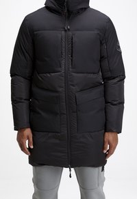 Veste matelassée noire avec capuche, fermeture zippée à l'avant et deux grandes poches. Tissu lisse et mat avec panneaux matelassés et logo embossé.
