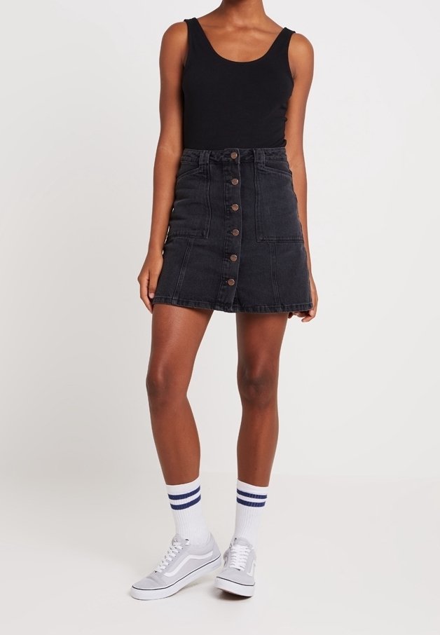 Femme portant un haut noir sans manches, une jupe en denim noire à boutons sur le devant, des baskets blanches et des chaussettes blanches avec deux bandes bleues.