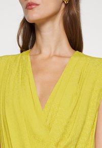 Robe jaune avec un décolleté en V profond, présentant un motif subtil et un tissu léger. Accentuée par un détail texturé à l'épaule.