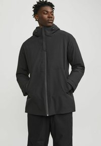 Chaqueta negra impermeable con capucha, cierre de cremallera y dos bolsillos laterales; presenta una textura suave y mate y un diseño minimalista.
