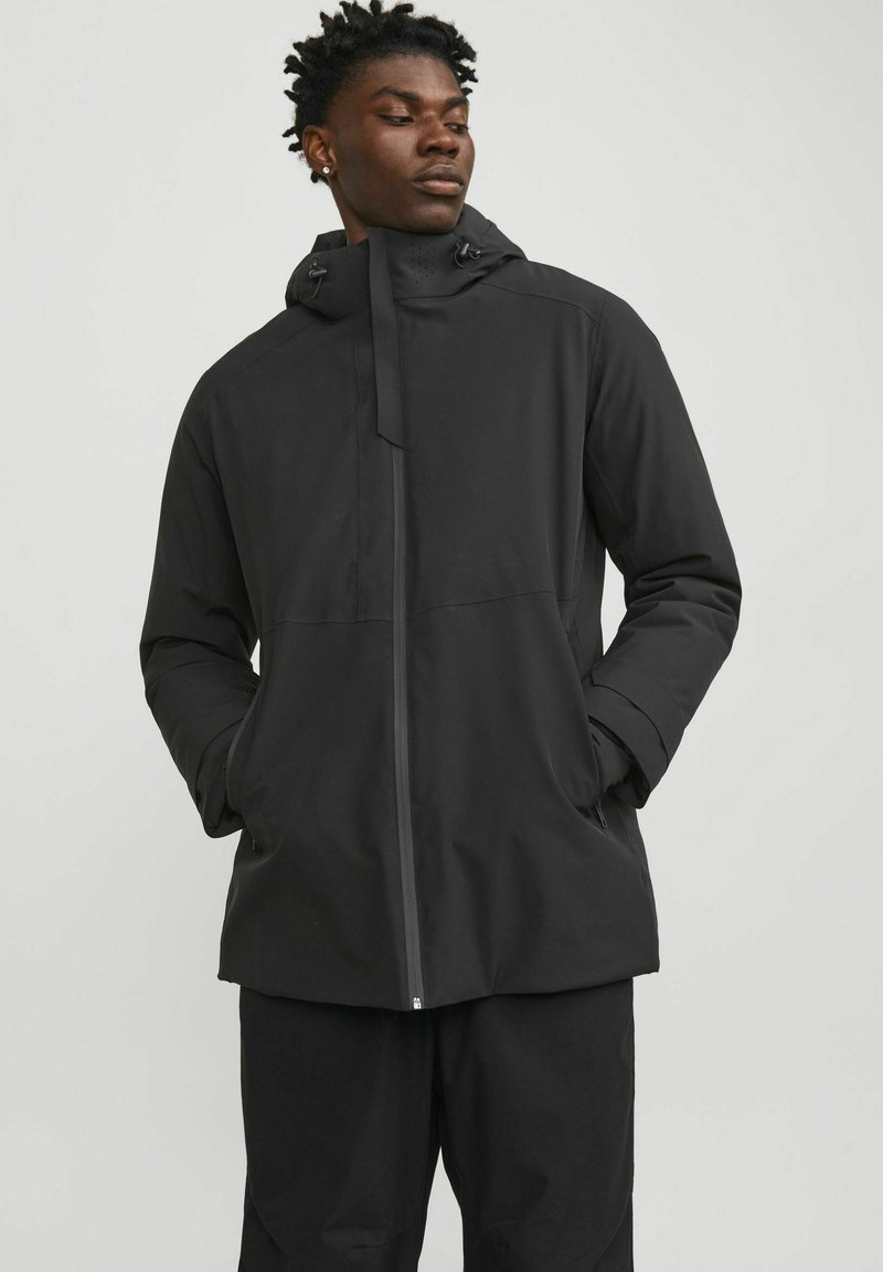 Chaqueta negra impermeable con capucha, cierre de cremallera y dos bolsillos laterales; presenta una textura suave y mate y un diseño minimalista.