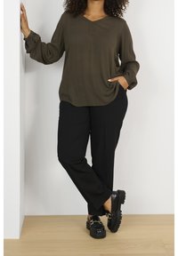 Femme appuyée contre un mur blanc, portant un blouse vert olive, un pantalon noir, des mocassins noirs, avec une main dans la poche et l'autre reposant sur le mur.