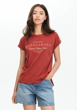 Joven mujer con una camiseta roja de Copacabana y shorts vaqueros azules, de pie con una mano en el bolsillo sobre un fondo blanco.