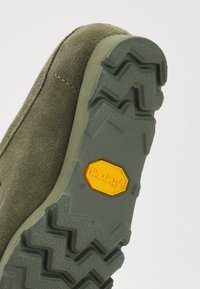 Semelle de chaussure de randonnée en daim vert avec un motif de crampons en zigzag. Présente un logo Vibram jaune, offrant durabilité et traction pour une utilisation en extérieur.