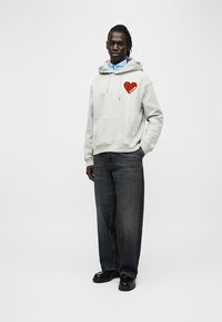 Sweat à capuche gris avec un motif de cœur rouge sur la poitrine, associé à un jean foncé et des chaussures noires. Le modèle se tient devant un fond blanc uni.