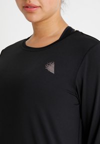 Crna sportska majica dugih rukava s okruglim izrezom, glatke teksture i malim geometrijskim logom svijetlosive boje na području prsiju.