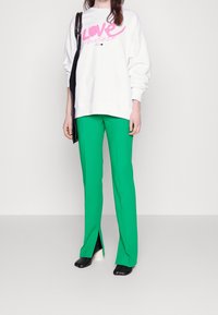 Sudadera blanca con el texto "QUIÉRETE" en rosa, combinada con pantalones ajustados verdes brillantes con aberturas laterales y zapatos negros con tacones blancos.