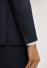 Manche d'une veste de costume bleu foncé à carreaux avec quatre boutons noirs, manchette de chemise blanche visible au-dessus de la main sur le côté.