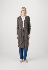 Cardigan longo de frente aberta em malha multicolorida com texturas variadas; combinado com jeans azul de cintura alta e sandálias de tiras.