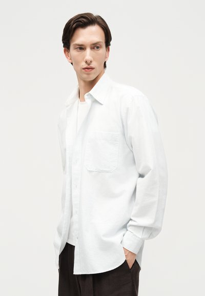 Νεαρός άνδρας με σκούρα μαλλιά, φορώντας λευκό πουκάμισο με μακριά μανίκια και σκούρο παντελόνι, στέκεται με το ένα χέρι στην τσέπη, κοιτώντας προς τα αριστερά.
